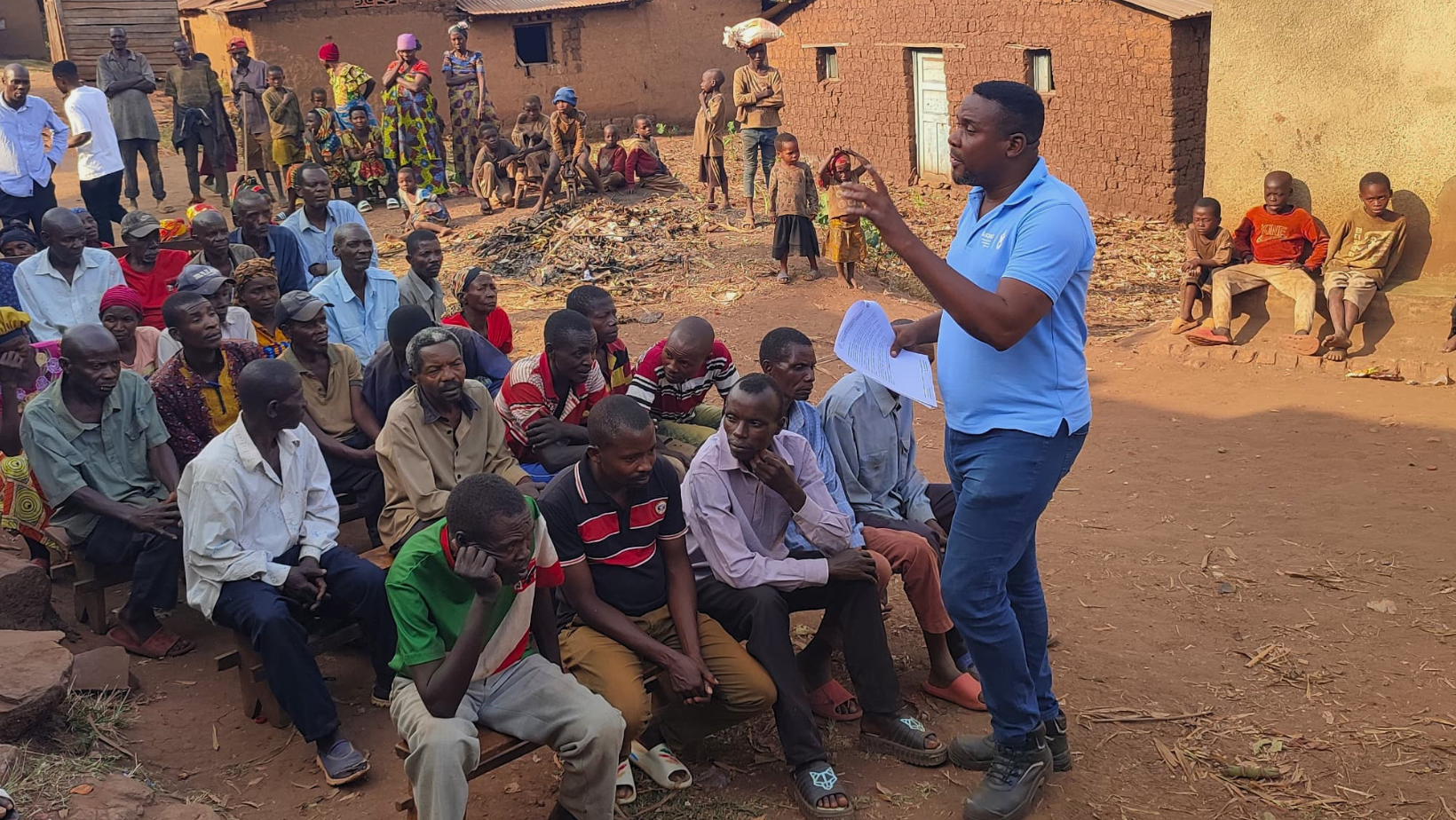 Un operatore in maglia azzurra parla con fogli in mano a una comunità locale seduta all'aperto durante un incontro formativo sul progetto idrico in Burundi.