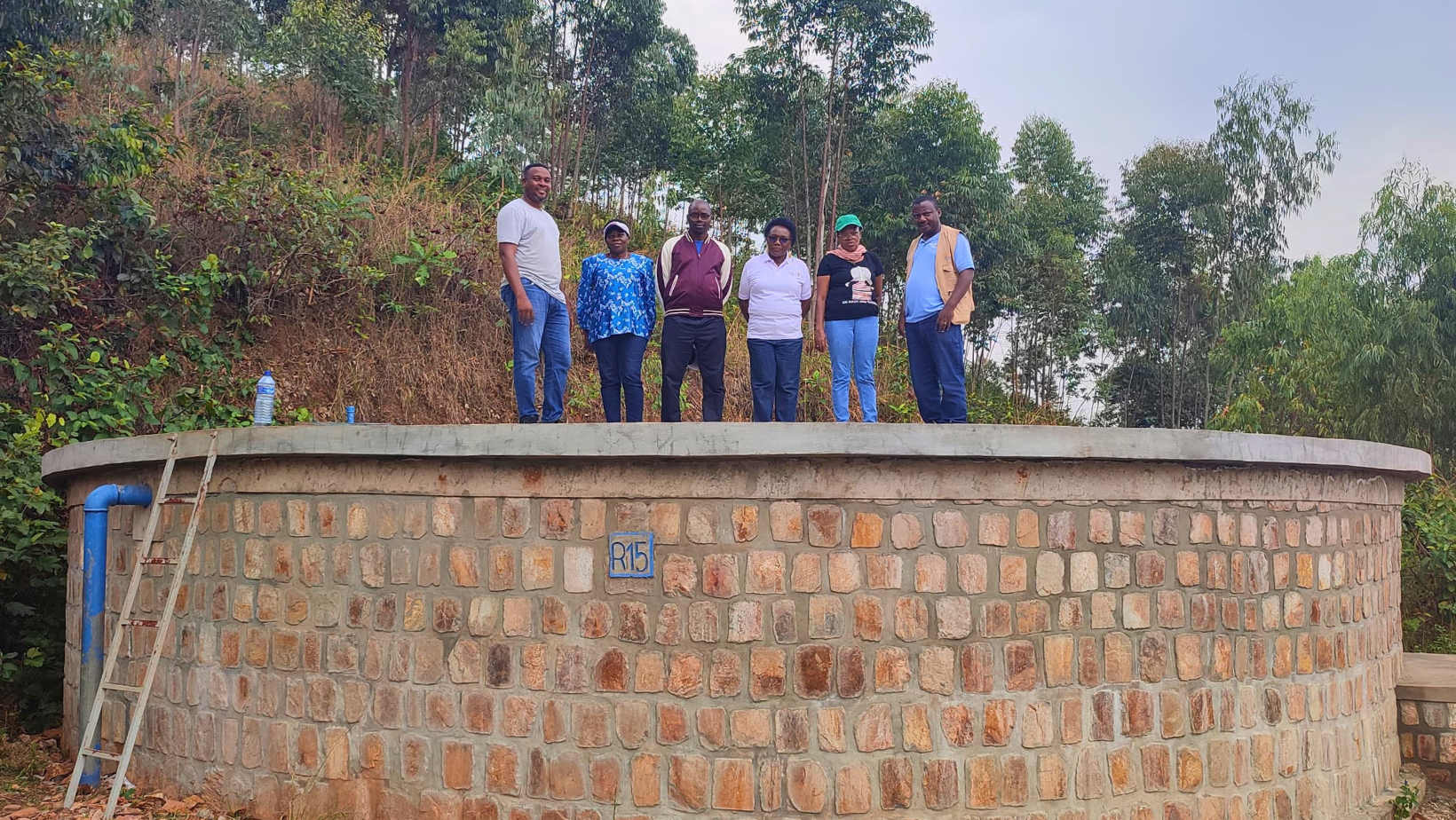 Sei persone, quattro uomini e due donne, in posa su un grande serbatoio d'acqua in muratura con la scritta "R15", in un ambiente rurale con vegetazione fitta in Burundi.