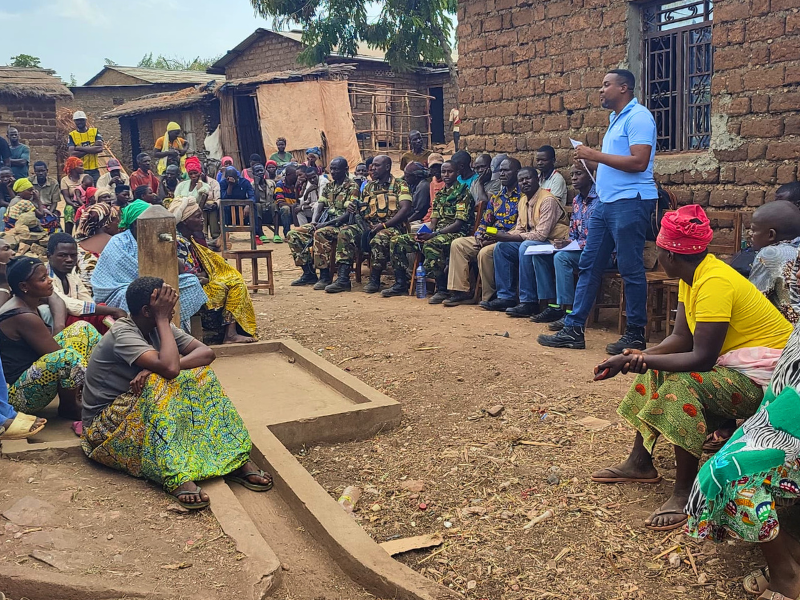 Incontro formativo a Cibitoke, Burundi: operatore AMU parla alla comunità locale seduta all'aperto vicino a una fontana.