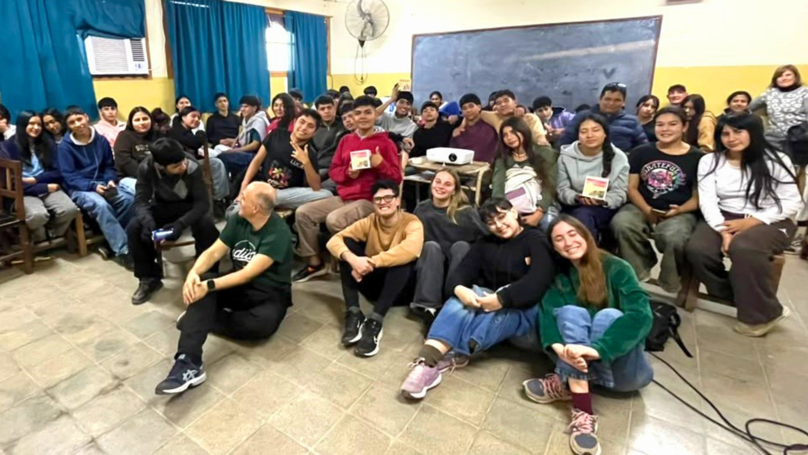 Un numeroso gruppo di giovani studenti argentini seduti in un'aula con insegnanti, alcuni sul pavimento e altri sulle panche, che sorridono per una foto di gruppo del progetto Living Peace. La stanza ha pareti gialle, tende blu e una lavagna.