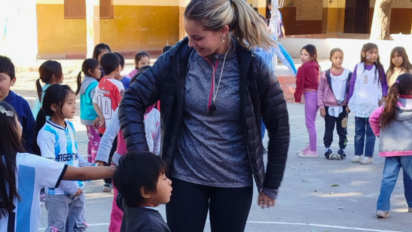 In una foto scattata in Argentina per la missione "Living Peace", una sorridente volontaria bionda, con una coda di cavallo e una giacca a vento nera su una maglia grigia, si china per interagire con un bambino piccolo, con la mano posata sulla sua testa. Il bambino guarda verso l'alto con un'espressione neutra. Attorno a loro c'è una folla di altri bambini e bambine che sorridono, alcuni con maglie dell'Argentina, su uno sfondo di un cortile scolastico con edifici gialli e un grande albero.
