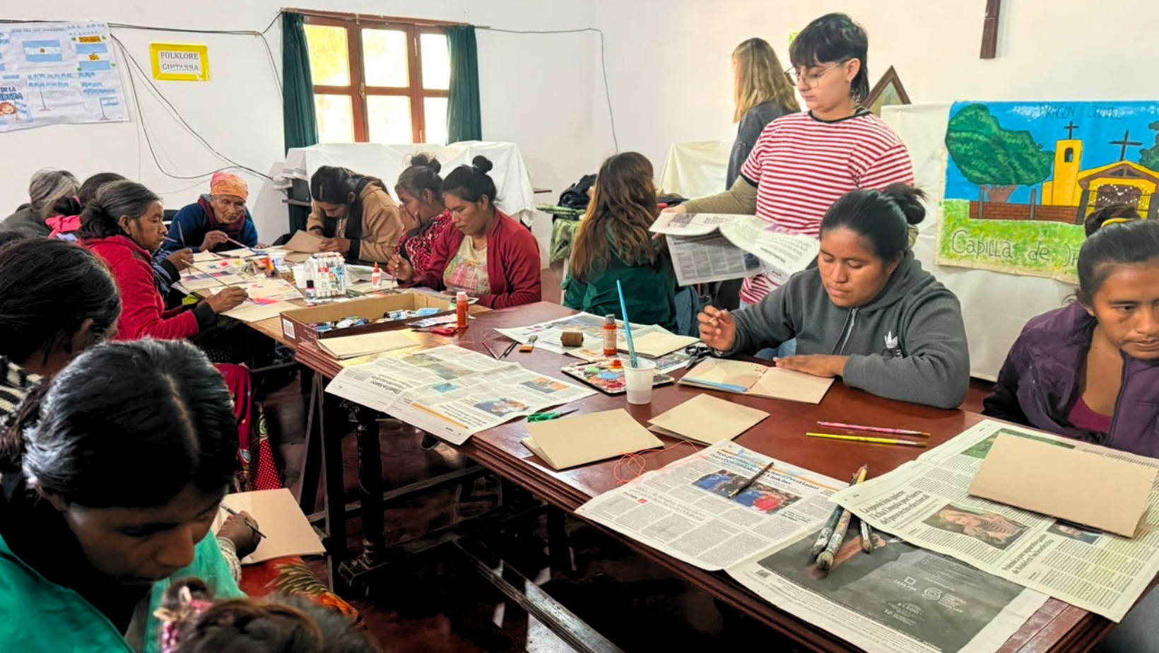 Un gruppo di donne indigene Wichí sedute attorno a grandi tavoli di legno in un'aula luminosa, partecipando a un laboratorio creativo. Sono concentrate sul dipingere e decorare quaderni artigianali con pennelli e colori. Sullo sfondo, due giovani volontarie di "Living Peace" e "Milonga", una con maglietta a righe, le assistono, mentre sulle pareti ci sono poster educativi e una bandiera argentina.