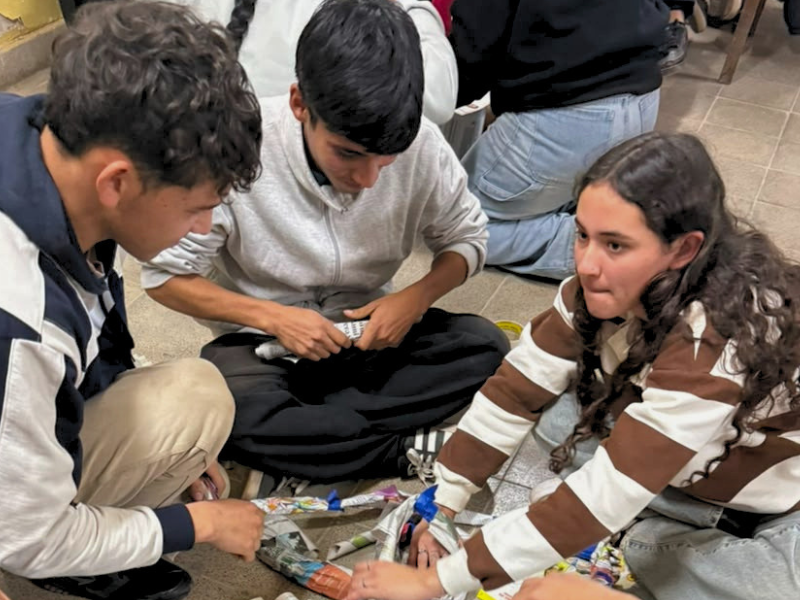 Tre Giovani Ambasciatori di Pace partecipano a un laboratorio sulla leadership positiva in una scuola argentina, collaborando seduti a terra per assemblare una struttura di carta di giornale con nastro adesivo.