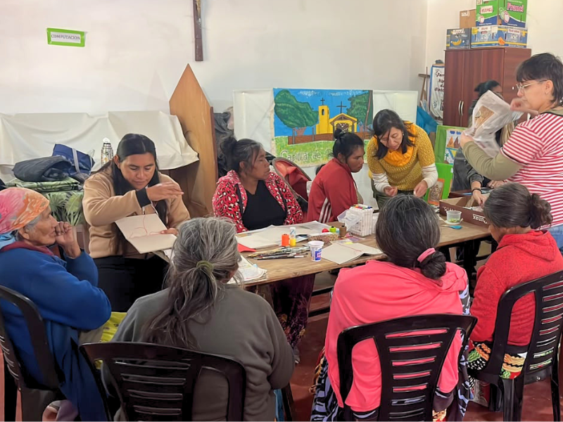 Un gruppo di donne indigene Wichí sedute attorno a grandi tavoli di legno in un'aula luminosa, partecipando a un laboratorio creativo