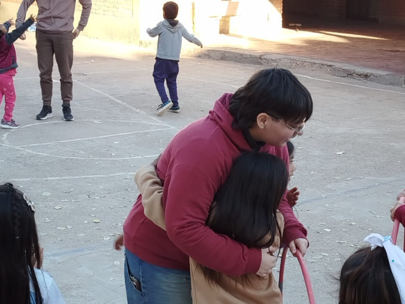 In primo piano, una giovane volontaria con gli occhiali e una felpa bordeaux abbraccia calorosamente una bambina dai lunghi capelli scuri nel cortile di una scuola. Sullo sfondo si vede un piazzale di cemento all'aperto con linee tracciate a terra, dove altri bambini e un adulto giocano e si muovono liberamente durante le attività della missione Living Peace in Argentina.