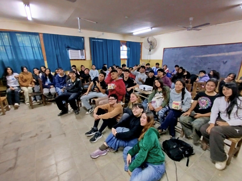 Un numeroso gruppo di giovani studenti argentini seduti in un'aula con insegnanti, alcuni sul pavimento e altri sulle panche, che sorridono per una foto di gruppo del progetto Living Peace. La stanza ha pareti gialle, tende blu e una lavagna.
