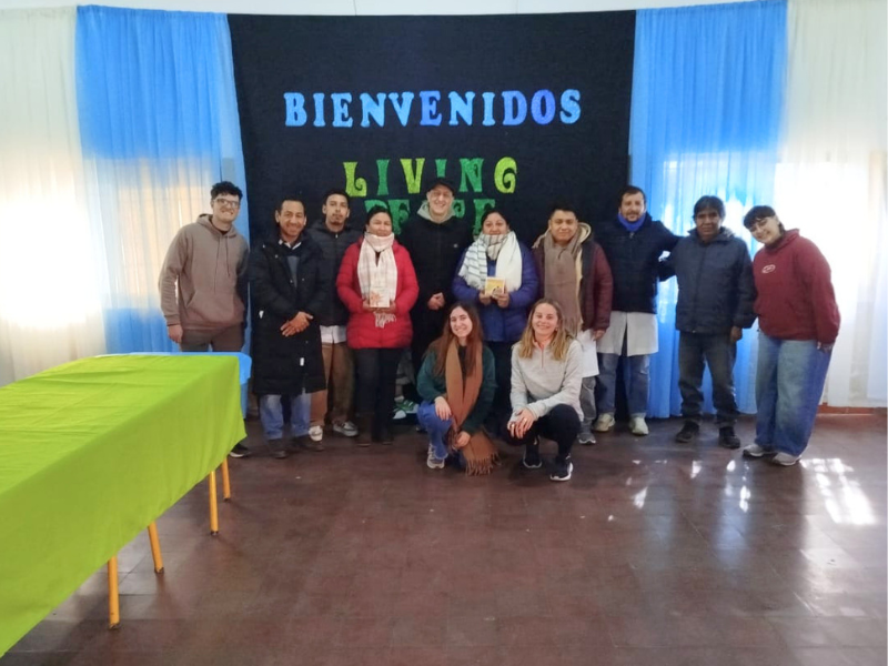 Foto di gruppo all'interno di una sala con undici persone, tra cui i giovani volontari e alcuni adulti della comunità locale. Sullo sfondo è appeso un grande pannello nero con le scritte colorate "BIENVENIDOS" in azzurro e "LIVING PEACE" in verde, incorniciato da tende bianche e azzurre. Sulla sinistra si nota un lungo tavolo coperto da una tovaglia verde acceso. Due ragazze sono accovacciate in primo piano al centro, mentre gli altri sono in piedi, tutti sorridenti e con abiti invernali.