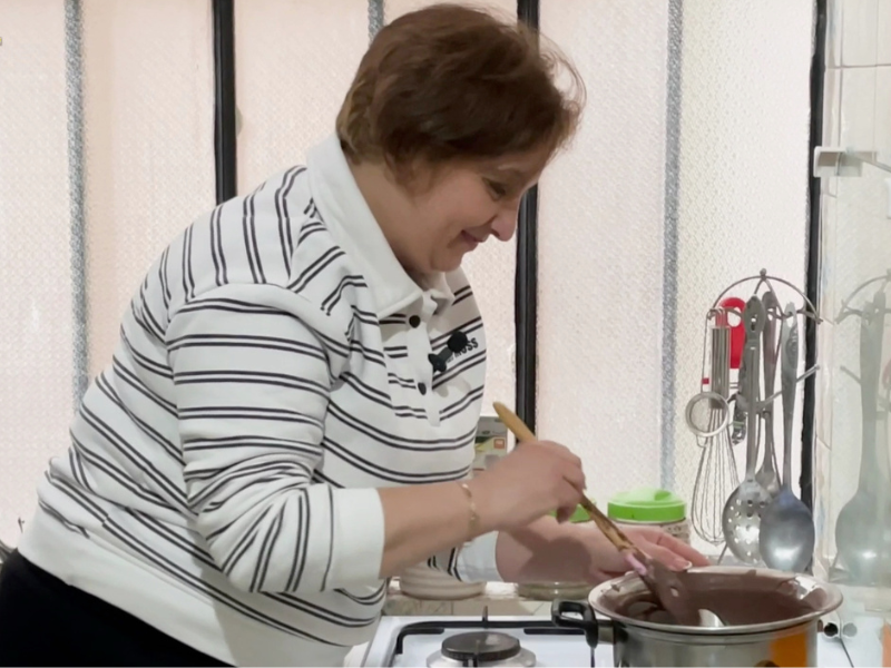 Shireen, sorridente, mescola il cioccolato fuso in una pentola con una spatola di legno in una cucina. Indossa una felpa a righe bianche e nere.