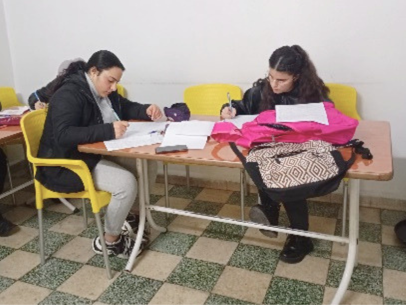 Due studentesse concentrate a studiare e scrivere a un tavolo con due zaini colorati.