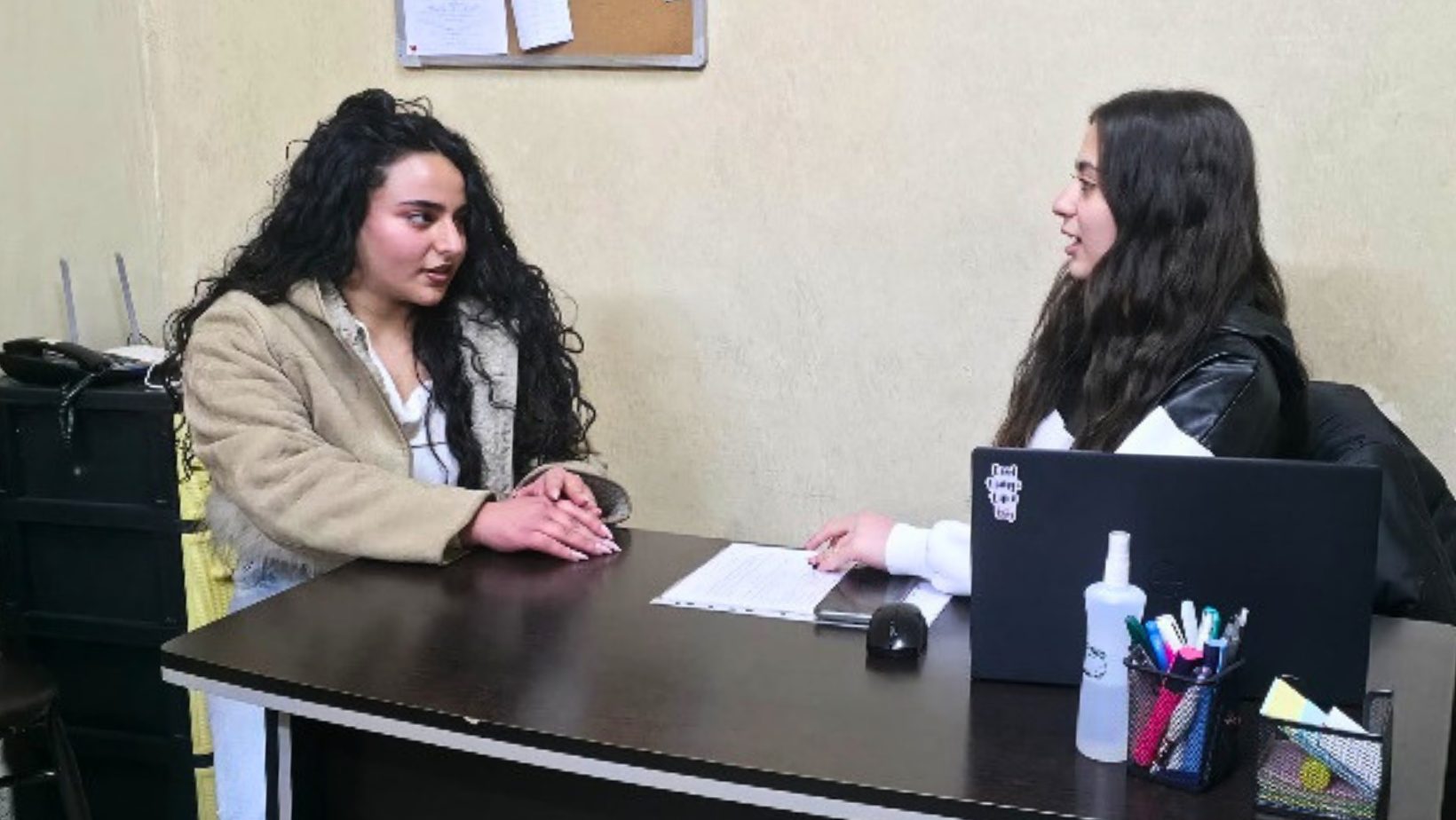 Due giovani donne sedute a una scrivania in un ufficio semplice, una a sinistra con una giacca beige che guarda l'altra a destra mentre parla. Una bacheca di sughero con fogli appuntati è visibile sullo sfondo.