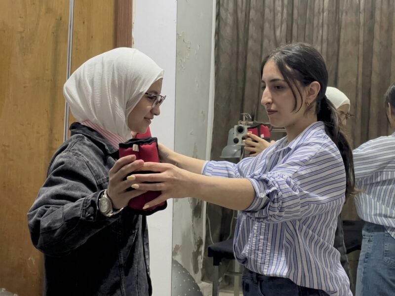 Una terapista in camicia a righe azzurre guida una ragazza con hijab bianco e giacca di jeans scura durante un esercizio di fisioterapia. Entrambe tengono con le mani un piccolo peso o supporto di colore rosso.