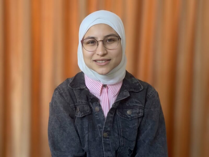 Un ritratto fotografico a mezzo busto di una giovane donna sorridente. Indossa un hijab bianco pulito, occhiali con montatura sottile a filo, una camicia a righe sottili rosa e bianche e una giacca di jeans scura slavata. È posizionata di fronte a un background a tinta unita, sfocato e caldo, color albicocca o pesca.