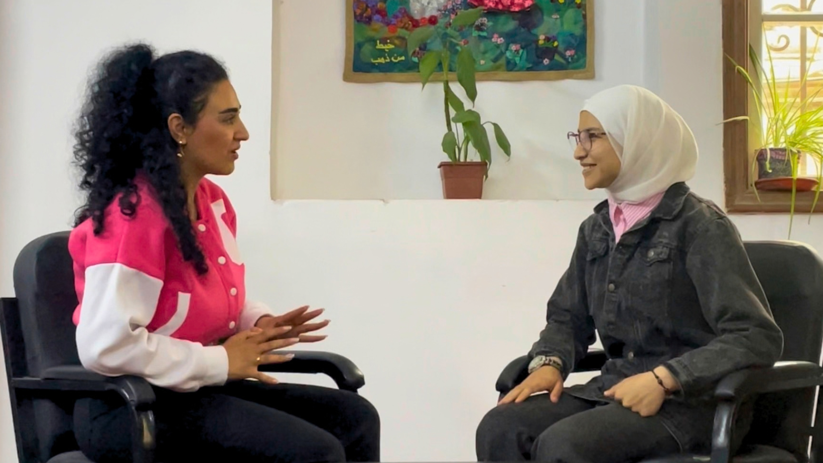 Due donne sedute di fronte durante un'intervista. Una donna a sinistra indossa una giacca rosa e bianca, una a destra indossa un hijab bianco e una giacca di jeans e sorride. Lo sfondo ha pareti bianche, piante e un arazzo appeso.
